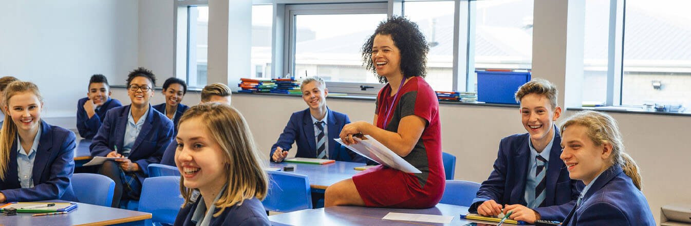 Teacher sitting on desk amongst secondary school students, all smiling to the front of the classroom