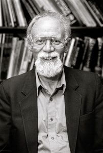 Black and white portrait photograph of Eric Jones. He is looking to camera, in front of bookcase.
