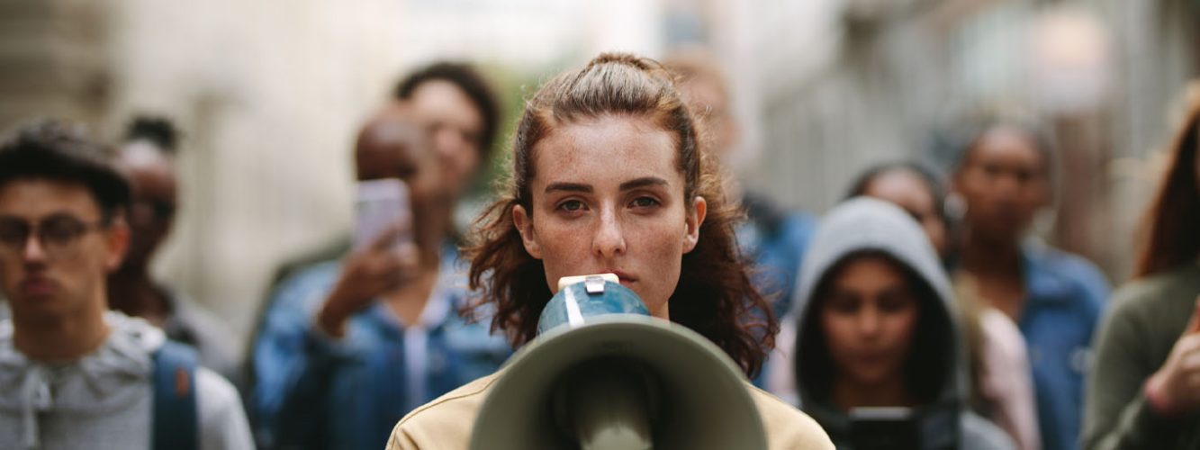 Protest scene, woman in foreground with megaphone, with crowd of people out of focus behind her