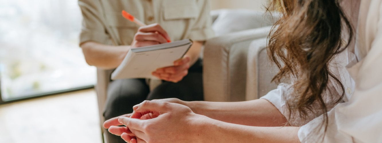 Woman's hands held together, sitting towards person taking notes on clipboard in therapy session, signifying skills needs for practitioners in complex trauma