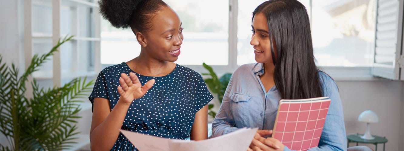 Two women in discussion in mentorship office environment, holding notebooks and paper
