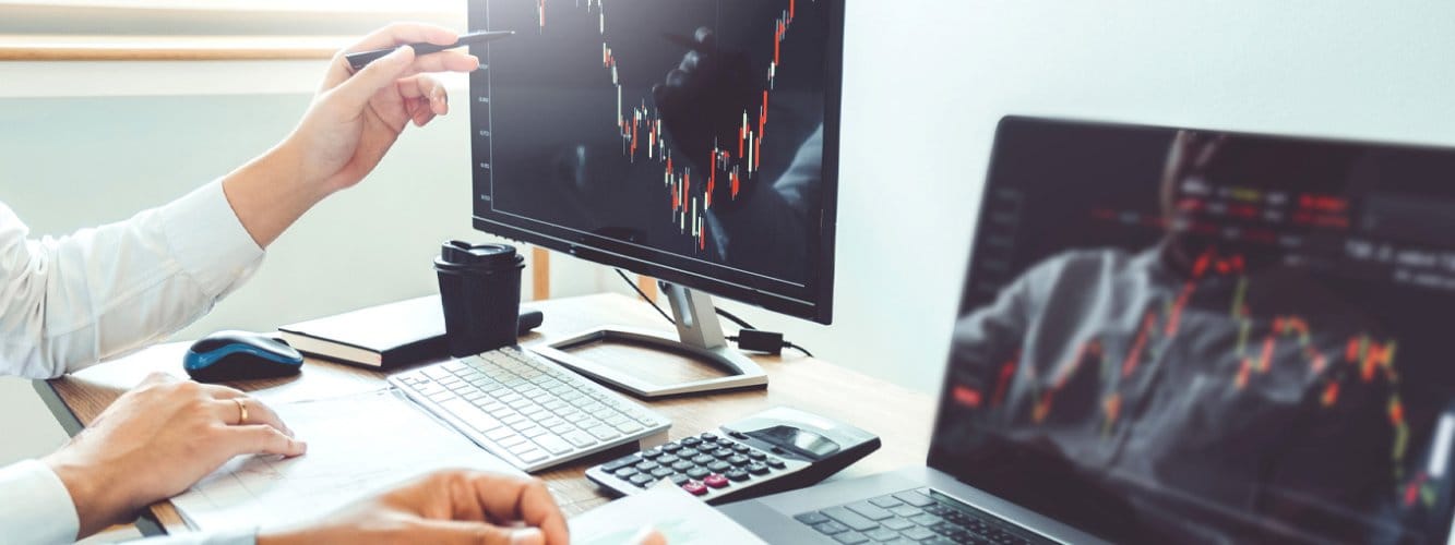 Two computer screens showing market trends on graphs, two people at desk, one pointing to screen