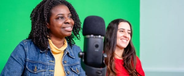 Two female students in digital studio, with green screen behind them and podcast-style microphone in front of them