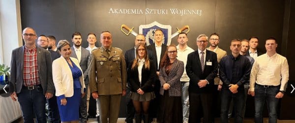 Large group of people in front of crest with sign: Akademia Sztuki Wojennej (War Studies University).