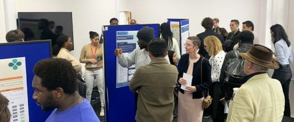Room split down middle with presentation boards, showing various posters. Students and academics are viewing the various posters.