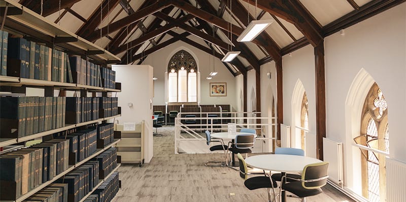 The Franciscan library: Tall shelves of books on the left, working spaces throughout, all within a building with historic and modern features including arched windows, and a high, vaulted ceiling 